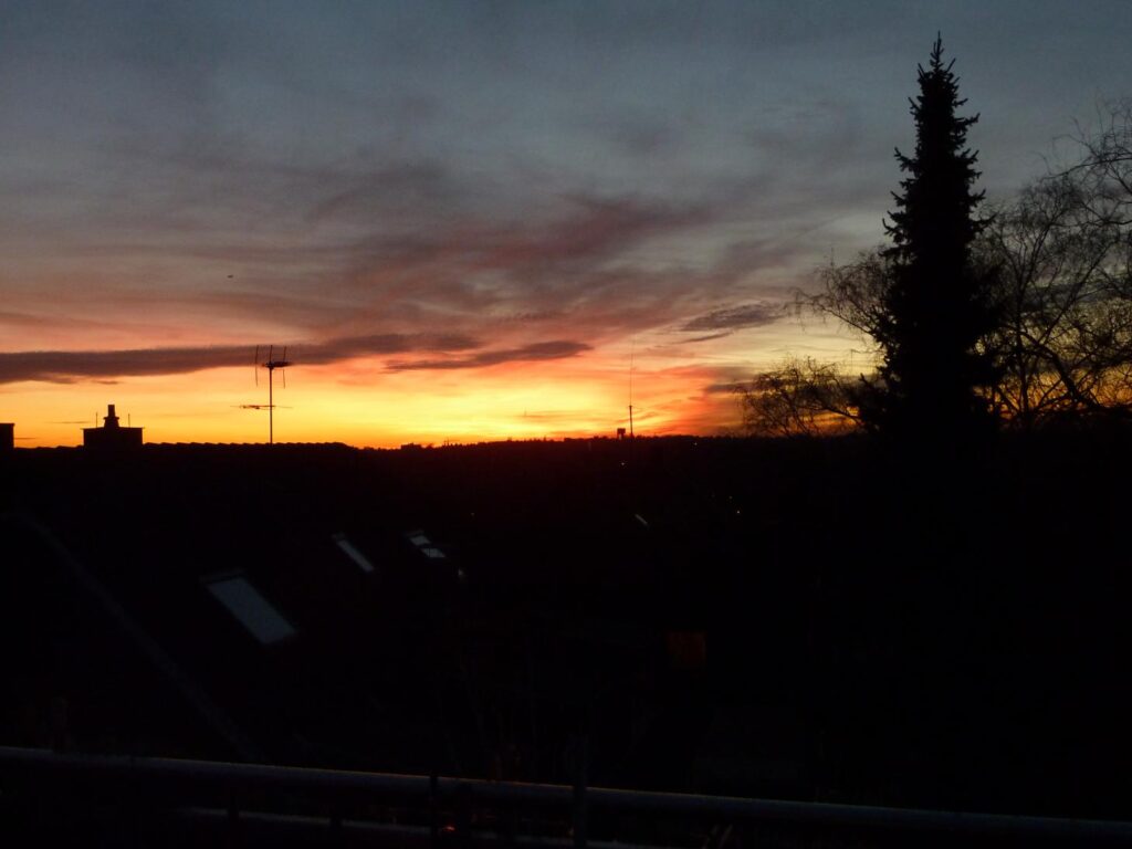 Abendstimmung. Im unteren Drittel des Bildes liegen Hausdächer und Gärten bereits im Dunkel. Rechts zeichnen sich Bäume - eine Fichte und eine herbstliche entblätterte Birke als schwarze Shilouetten vor dem Nachthimmel ab. Am Horizont noch ein Streifen in Gold und Rot, darüber vioellett Wolken am rauchfarbenen Abendhimmel. Gleich wird das Dunkel der Nacht herrschen.