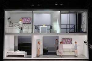 Ein weißes, kastiges Puppenhaus ist Thilos Reuthers Bühne, indem sich auf zwei Stockwerken die Handlung abspielt.