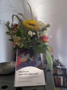 Ein leicht verwelkter Spätsommerblumenstrauß mit Sonnenblume, an dessen Vase ein Buch lehnt: "Was bin ich, wenn alle anders sind?" ist der Titel