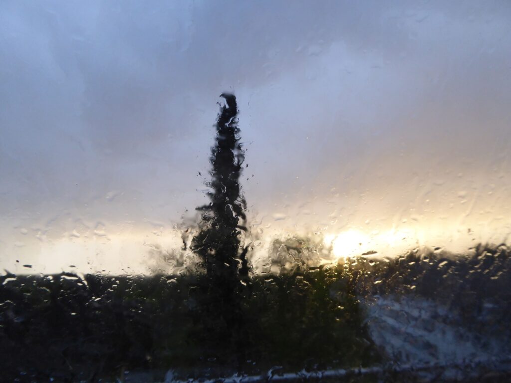 Die Sonne kurz vorm Abendrot tief an einem aufreißenden Himmel gesehen durch ein Fenster voller Regentropfen, in denen sich das Abendlicht bricht