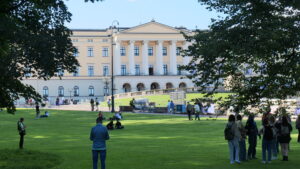 Das königliche Schloss in Oslo an einem sonnigen Tag mit zahlreichen Menschen auf der Wiese im Park davor.