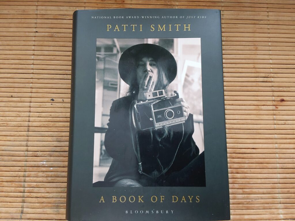 Patti Smith "A book Of Days", auf dessen Cover die Künstlerin mit einer Polraidkamera in der Hand zu sehen ist. Sie schlägt die freie Hand vor den Mund, als sei sie erstaunt oder als habe sie sich selbst dabei ertappt, wie sie sich selbst fotografiert.