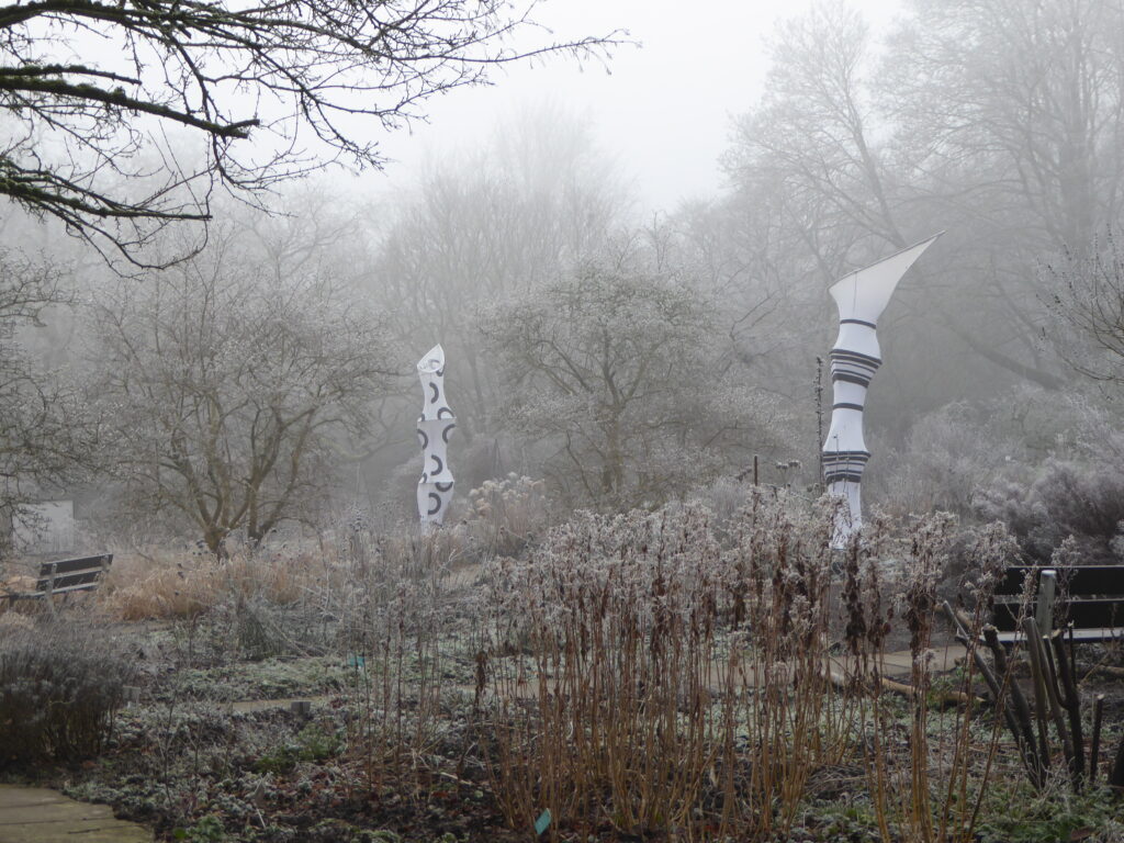 Zwei seltsam geformte Skulpturen mitten in einem Staudenareal im Nebel