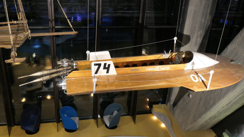 Ein Holzboot mit Motor schwebt vor der dunklen Fensterfläche. Dass es ein größeres Boot sein muss, ist leicht daran zu erkennen, dass eine Eben unter ihm mehrere große Sessel und andere Objekte Platz finden.