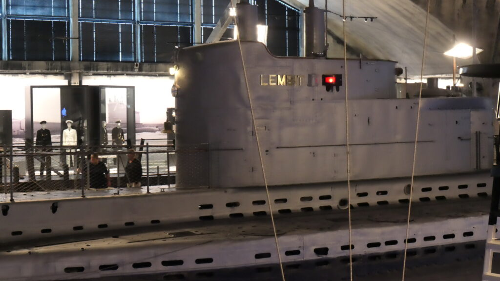 Das U-Boot, die 1937 gebaute Lembit, etwas näher vpn der Seite betrachtet. Auf der Plattform neben dem Schiff bzw. aus dieser Perspektive im Hintergrund des Bildes, zeigen Schaufensterpuppen elegante Uniformen.