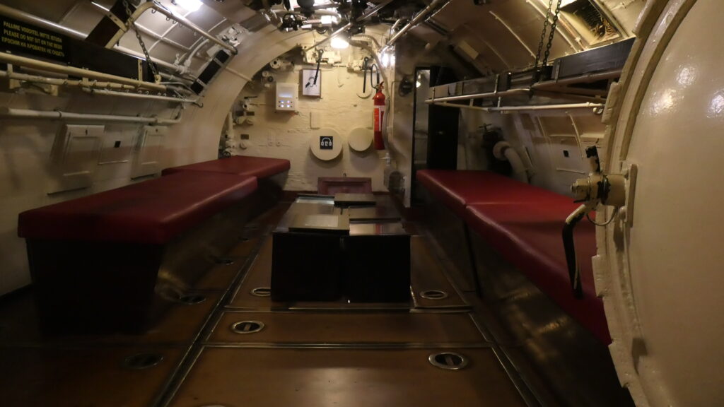 Hängende Betten über Kästen mit roten Matratzen auf beiden Seiten des Rumpfes. Im Vordergrund ist rechts angeschnitten das Ende eines (Torpedo)Rohrees zu sehen. Da hier die Wände verkleidet sind und das Licht ein warmes ist, wirkt dieser Teil des U-Bootes geradezu heimelig.