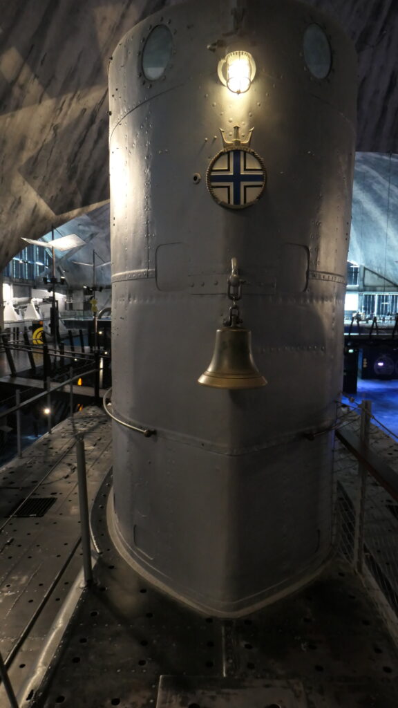 Frontal und recht nah aufgenommen, der Turm der Lembitu. Über der Messingglocke ein Kreis mit einem schwarzen, gerahmten Kreuz auf weißem Grund. Der Kreis mit seinem dreizackigen Krönchen wird von einer Lampe am aberen Ende des Turmes erleuchtet. An den Seiten desselben geht der Blick in die blaue Tiefe des Halle.