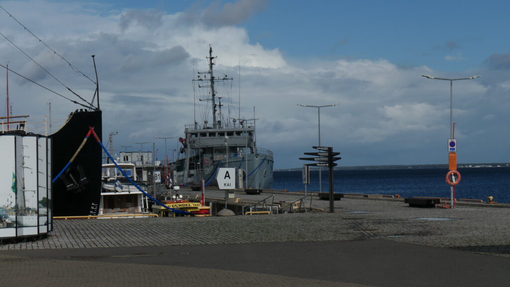 Blick vom Wasserflugzeughafen hinaus auf den Pier. Links ragt noch gerade so der schwarze Bug der Suur Toll ins Bild, rechts ein Stück den Kai hinunter liegt die Valvas. Das Meer zu ihrer rechten ist dunkelblau. Über den Wolken am Horizont reißt der Himmel auf.