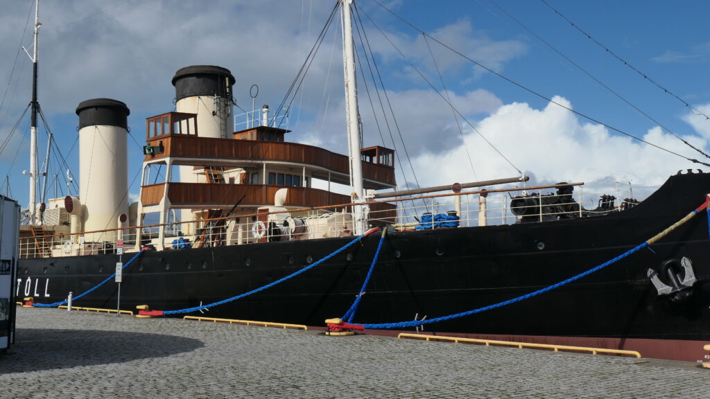 Die Suur Toll festgemacht am Wasserflughafen wirkt mit ihrem schwarzen Rumpf, den beiden weißen Schonsteinen mit schwarzer "Krone" unde dem mit rötlichen Holz verkleideten Aufbauh zwischen Schornsteinen und Mast in der Bootsmitte, der Brücke und "Ausgucke" enthält, recht imposant. Das verstärken auch die Wolken nebst blauen Himmelslücken darüber.