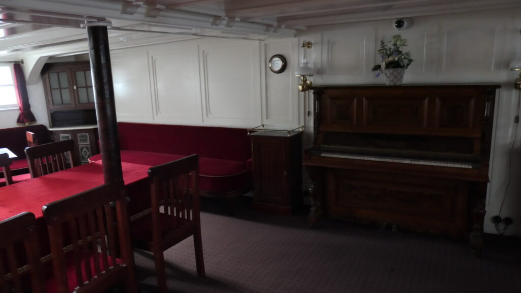 Die Messe der Suur Toll mit eleganten Holzstühlen und Bänken, rotplüschig bezogen, und einem Klavier samt Topfpflanze - ein einladender, wohnlicher Ort.