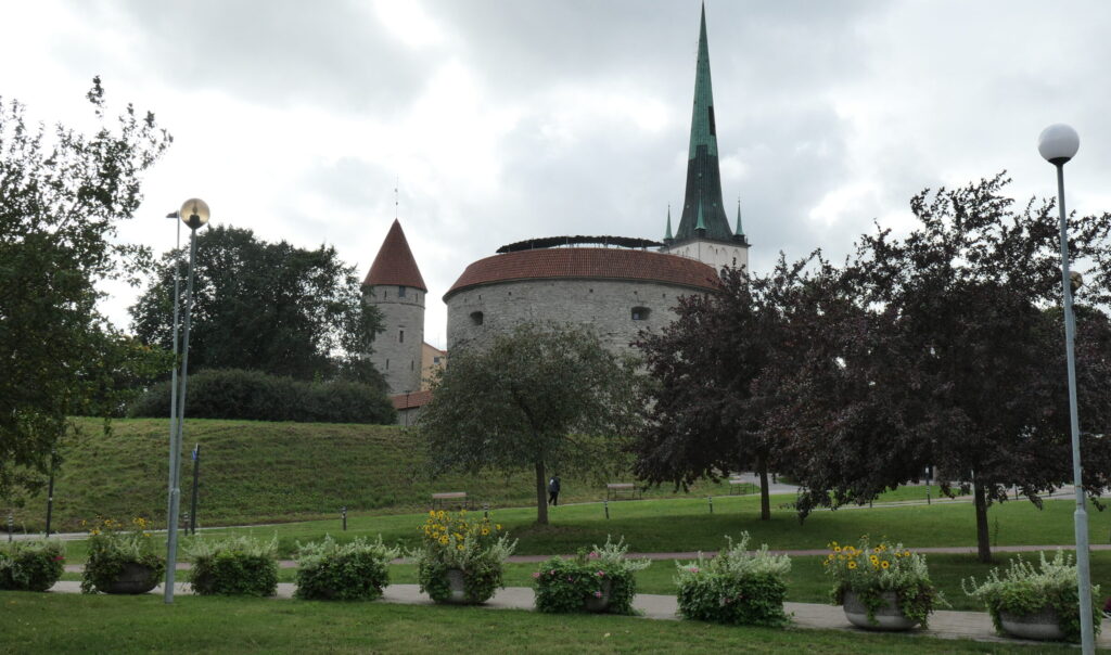 Die dicke Margarethe, ein sehr breiter mittelalterlicher Rundturm, leigt auf einem kleinen Hügel hinter einer Grünanlage und wird teils von Bäumen im Vorderund verdeckt. Hinter ihr erhebt sich der markante, ausgesprochen spitze Turm von St. Olaf.