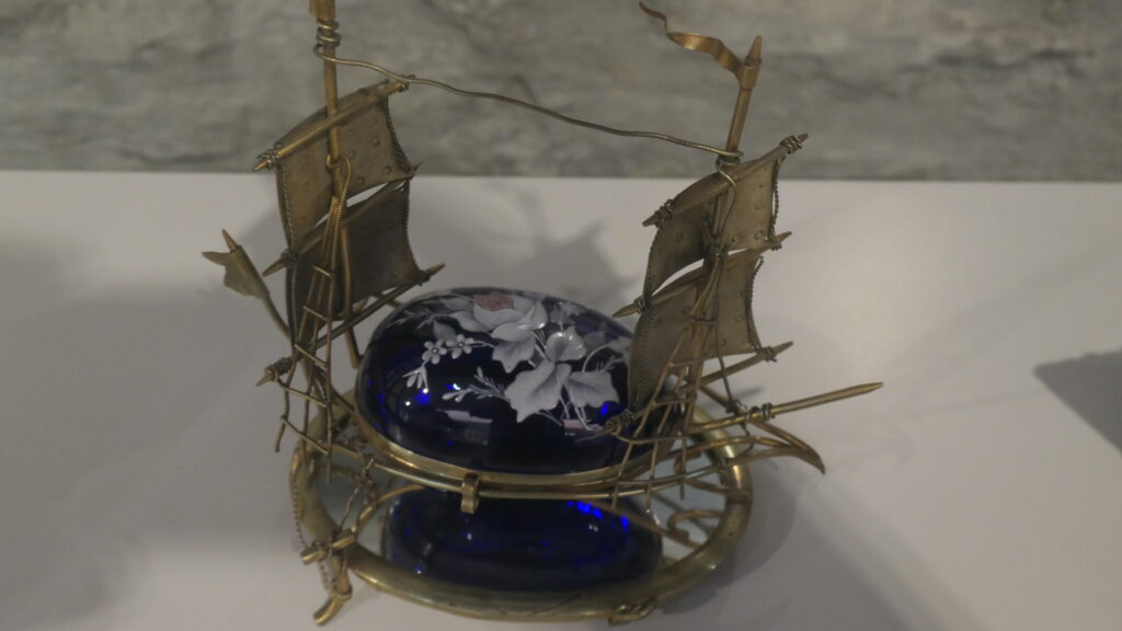 Ein blaues Fayence-Ei mit aufgemalter weißer Blume bildet den Rumpf des ungewöhnlichen Modellschiffes. Vor der Spitze und hinter dem stumpfen Ende des liegenden, mit Messing eingefassten Eis, bedindet sich jeweils ein Mast mit zwei Segeln. Hinten weht eine Fahne, vorn ragt der Bugspriet hervor.