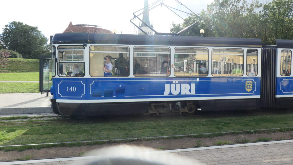 Eine alte, blaue Straßenbahn saust vorbei. "Jüri" ist weiß auf blauem Grund auf ihrer Seite zu lesen. Hinter der Tram sieht man noch das Dach der dicken Margarthe und den Turm von St. Olaf
