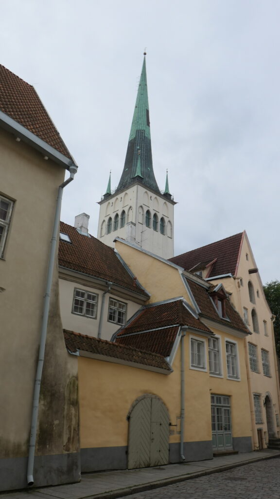 St. Olafs grünes Turmdach ragt in den rauen Himmel hinauf. Verwinkelte Altsadtbauten in unterschiedlichen Gelb- und Orangtönen säumen die Altstadtgasse, die den Blick auf die Kirche selbst versperrt.