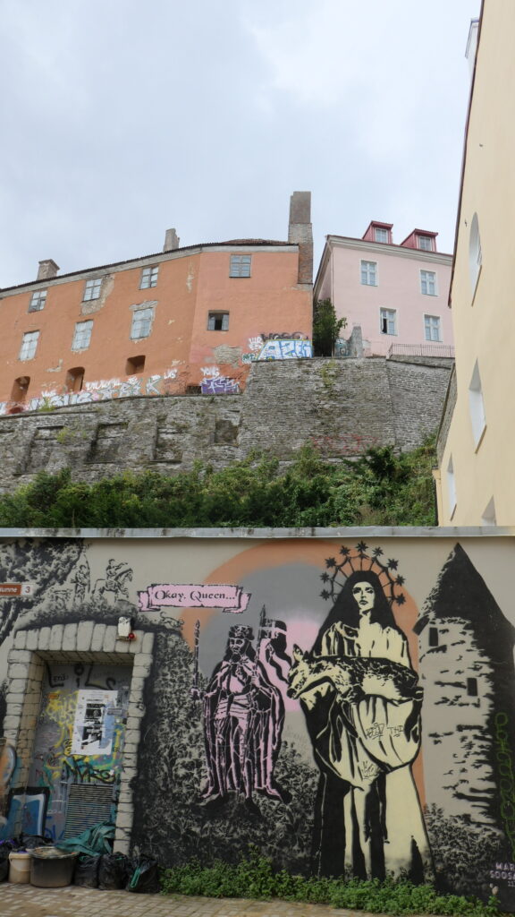 Blick von der Altstadt über die Stadtmauer hinauf zu mehrgeschossigen Gebäuden auf dm Dombet oberhalb der Stadtmauer. Auf der Eben des Betrachters ist eine weitere Mauer mit einm Durchgang. Auf dieser Mauer zwigt ein Mural eine Frau mit Krone oder strahlenbewehrtem Heiligenschein, die auf den Betrachter zugeht und ein totes Reh trägt. Links hinter ihr steht ein mittelalterlich gewandeter König, über dessen Kopf in einem Spruband steht "Okay Queen ...." Rechts neben ihr ist der Plate Tower zu erkennen.
