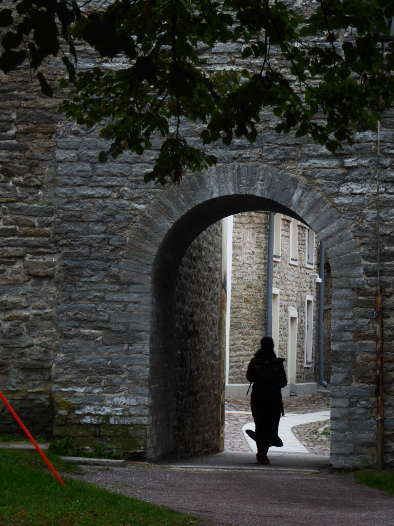 Blick aus dem Park auf einen der zahllosen Durchgänge in der Stadtmauer, die hinein in die Altstadt führen. Der Vordergrund ist recht dunkel, ddie Altstadt dahinter heller. Im durchgang die Shilouette einer Person mit weit schwingendem Mantel, die in die Altstadt geht.