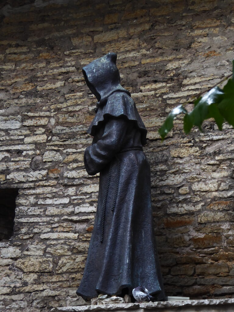 Vor der Stadtmauer beim Gartne des dänischen Königs wendet sich diese schwarze Skulptur eines Mönches bzw. seiner Gestalt verborgen im weiten Habit mit Kapuze, auf einem erhöhten Vorsprung nach links.