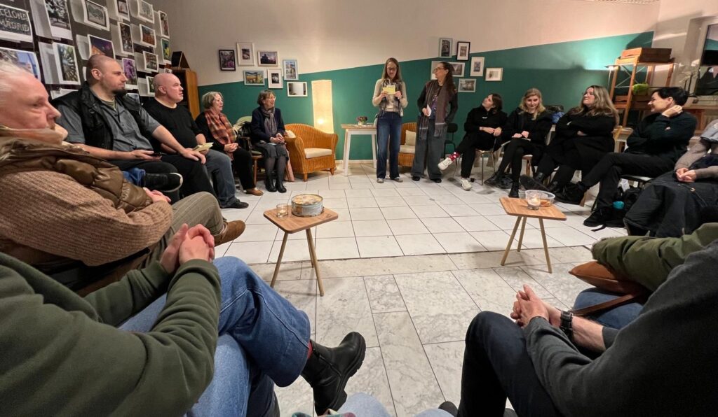 Das Publikum sitzt in eine großen Kreis. Im Vordergrund in der Mitte zwei Tischchen mit Keksen. Gegenüber am anderen Ende, sozusagen auf der Lesebühne, zwei Korbsessel und ein größerer Tisch, beleuchtet von einer großen Stehlampe. Davor stehen zwei Frauen mit Moderationszetteln.