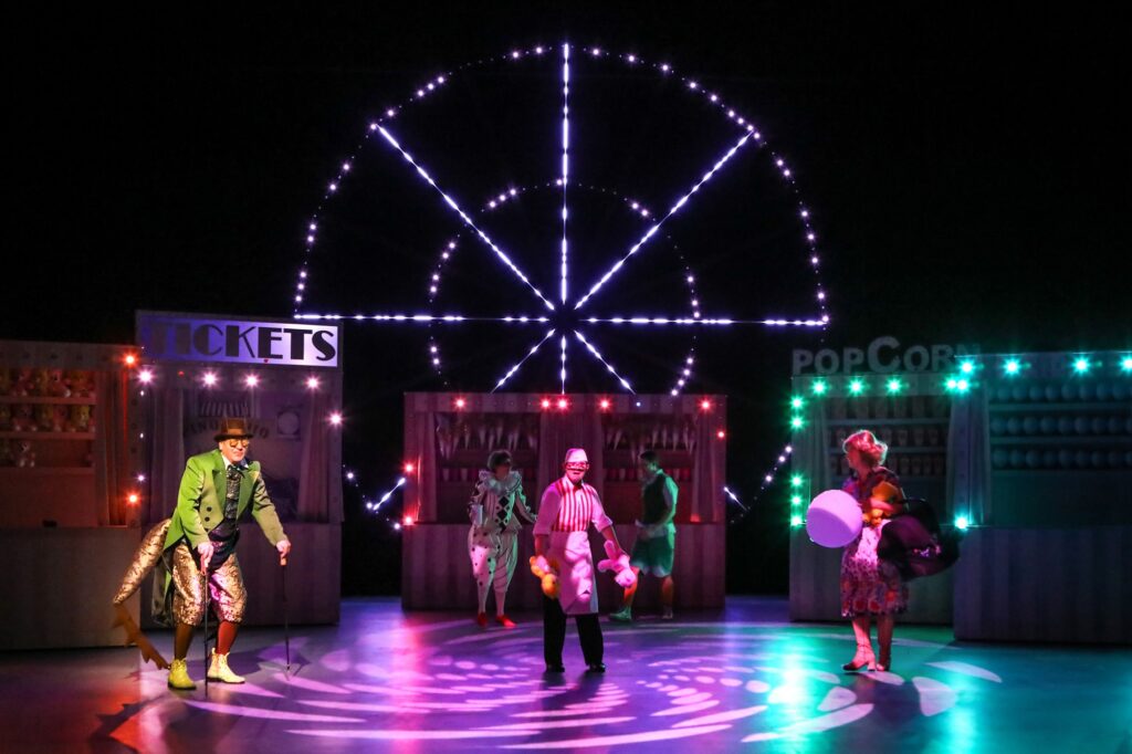 Die Magie eines Abends auf dem Jahrmarkt: lauter bunte Buden mit Losen, Popcorn, Teddybären und Wurfspielen vor einem leuchtenden Riesenrad. Auf dem Platz zwischen den Buden tanzt das Ensemble.