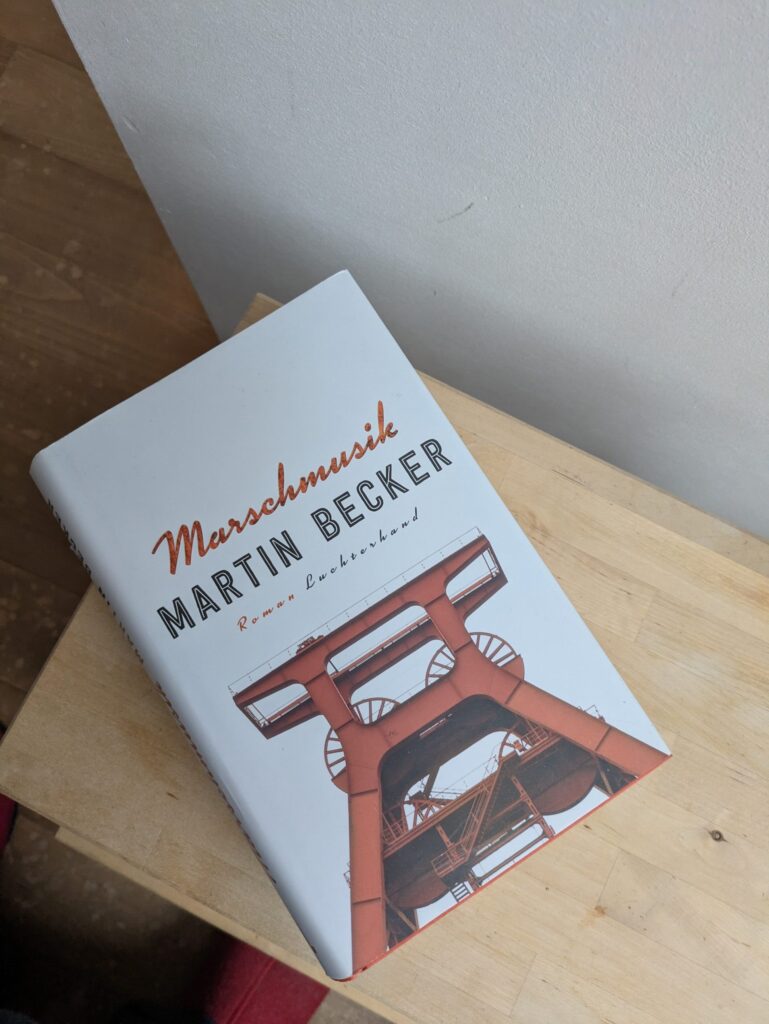 Auf einem Holzbrett liegt die Hardcoverausgabe von Martin Beckers Marschmusik. Der Hintergrund des Cover ist weiß, der Titel selbst stehr in roter, schräggestelter Kursvischrift über dem in Großbuchstaben (schwarz umrandet, innen leer) gesetzten Autreon Namen. Sehr viel kleiner, aber wieder kursiv steht darunter in Rot Roman und in Schwarz Luchterhand. Darunter ist freigetellt ein roter Förderturm aus Untersicht zu sehen.