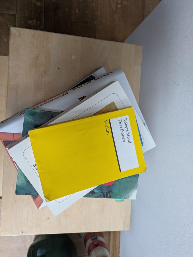 Ein Stapel Bücher (dieselben wie im bild zuvor) auf einem Holzbrett. Obenauf liegt die leuchtend gelbe Reclam-Ausgabe von Robert Musils "Drei Frauen"
