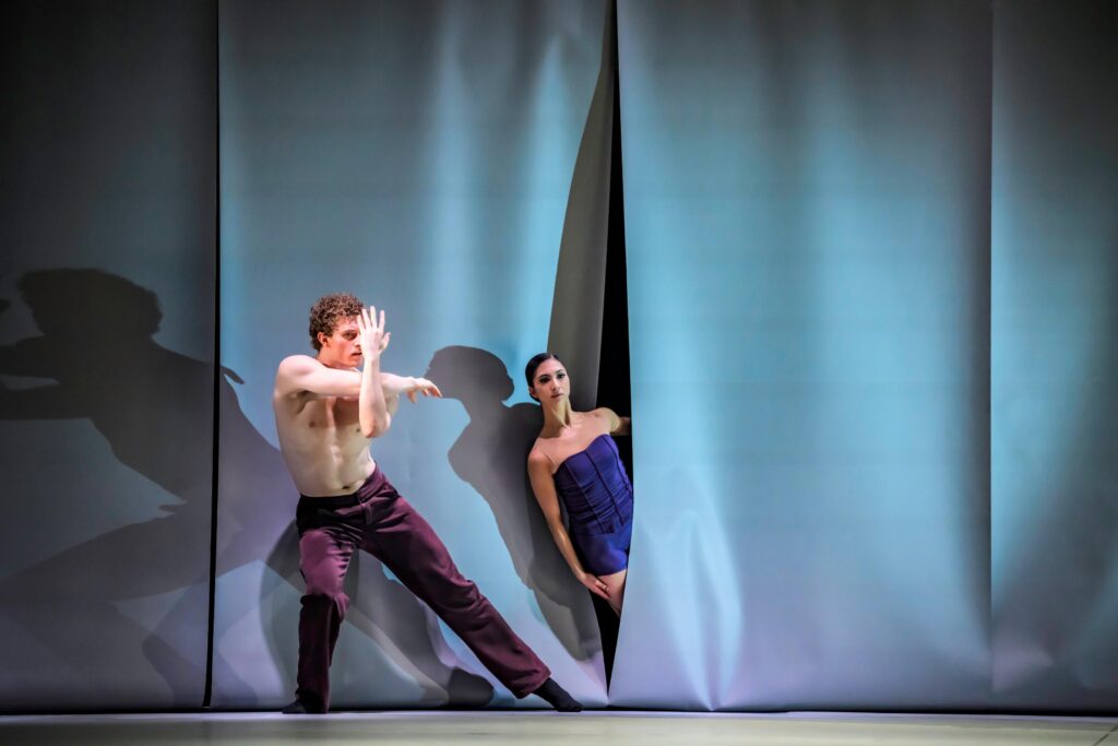 Vier weiße Rechtecke aufgespannt wie Segel, zwischen ihnen schlitzartige Öffnungen, beherrschen die Bühne. Ein Tänzer in purpurfarbener Hose neigt sich vor dem zweiten diagonal zur rechten Seite. Durch die Öffnung in der Mitte gleich neben ihm sieht man den Oberkörper einer Tänzerin in violetter Corsage, die sich mit angelehntem Arm parallel zu ihm neigt - eigentlich ein unmöglicher Winkel, sodass sie fallen müsste.