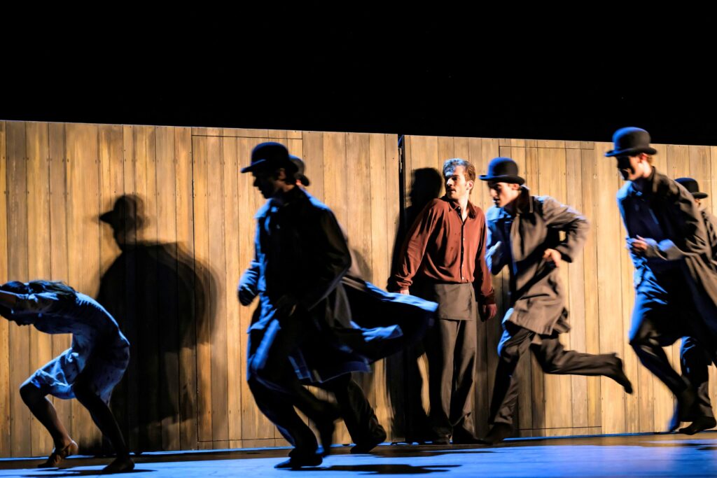 Ein Mann in Hemd und Hose steht im hellen Licht an eine Wand aus Holzbrettern gepresst da und schaut einer Gruppe von schattenhaften Männern mit Mänteln und Hüten nach, die hinter einer Frau in einem Kleid herlaufen.