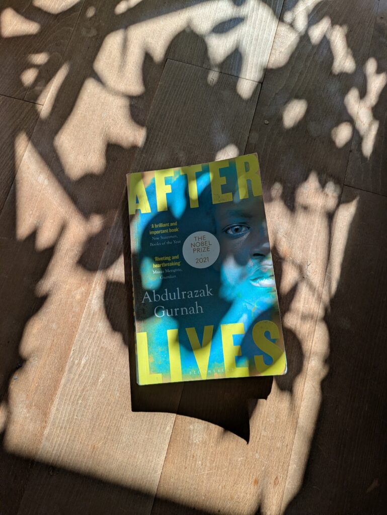 Auf einem sonnenbeschienen Fußboden, auf den ein Benjamini seinen Schatten wirft, liegt die Taschenbuchausgabe von Abdulrazak Gurnahs "After Lives". Der Titel prangt oben und unten in großen gelben Lettern auf gründem Grund, rechts am Rand angeschnitten das Gesicht eines mutmaßlich afrikanischen Kindes, das den Betrachter aufmerksam anblickt. Links in kleiner, gelber Schrift Kurzkritiken und direkt über dem "Lives" unten der Name des Autors. Doch vor allem der Blick des Kindes, das zwischen den beiden Worten des Titels hindurchblickt wie aus einem zu klein geratenen Fenster, fesselt.