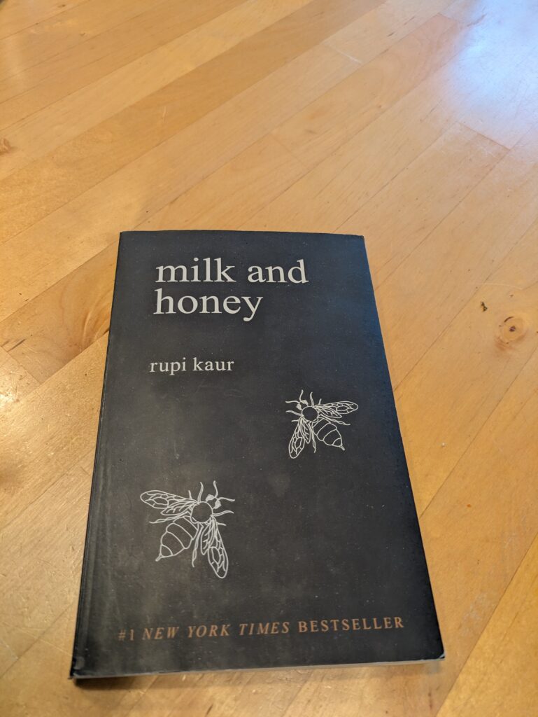 Auf dem schwarzen Cover von Rupi Kaurs "Milk und Honey", das auf einem hellen Holztisch liegt, steht oben der Buchtitel und darunter etwas kleiner der Name der Autorin. Zwei ebenfalls in weiß gezeichnete Bienen krabbeln Richtung Schrift - die eine aus der unteren linken Ecke, die andere weitere oben rechts. Ganz unten am Rand steht in dunkelgelber Schrift #1 New York Times Beststeller