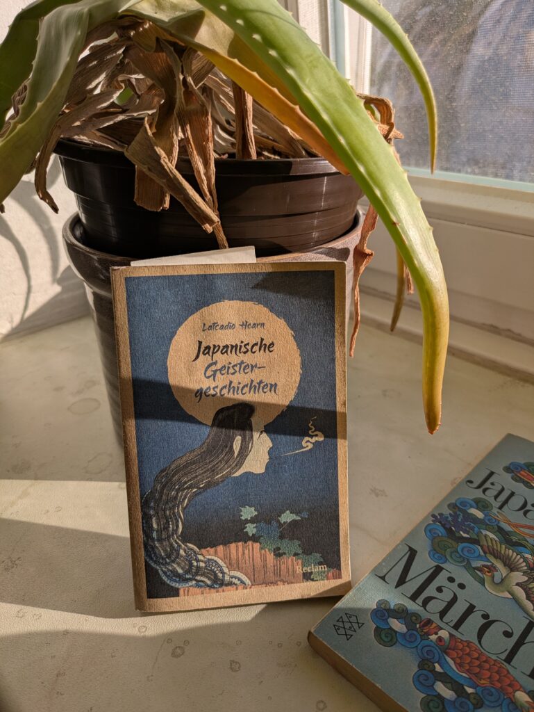 Auf einer Fensterbank lehnt das kleine Reclam-Büchlein am Topf einer aloevera. In der Bildmitte ziert eine Art stilisierter Vollmond den nachtblauen Himmel. In dem Rund steht in kleiner Schrift "Lafcadio Hearn" über dem größer gesetzten Titel "Japanische Geistergeschichten".In den Mond hinein ragt der menschliche Kopf eines Dämons oder Geistes, dessen schlangenartiger Körper sich aus der unteren Mitte über die linke Seite hinaufwindet. Er scheint aus einem hölzernen Gefäß zu kommen, an dem eine Pflanze mit efeuartigen Blättern entlangrankt. Das Gesicht des Wesens pustet sichtbar nach rechts etwas aus.