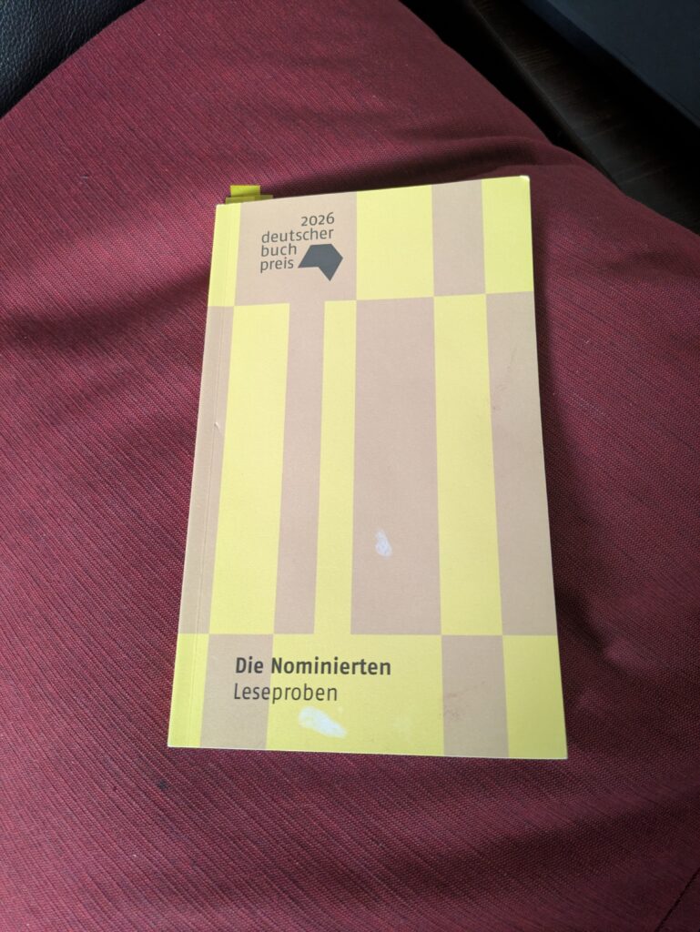 Auf dem länglichen, unregelmäßig gelb-braun gestreiften Cover des dünnen Buches steht oben links "2026 deutscher buchpreis" und linksbündig unten "Die Nominierten. Leseproben". Das leicht angeschlagene buch, aus dem oben kleine gelbe Klebenzettel als Markierung ragen, liegt auf einem dunkelroten Sofakissen.